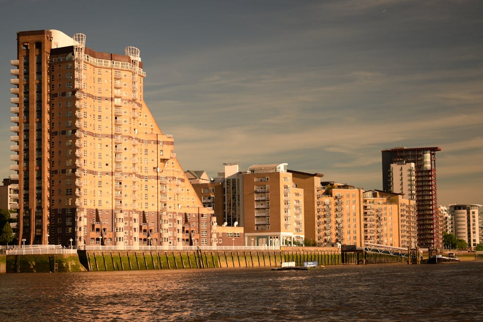 A row of modern multi-storey residential buildings along the riverbank in Chelsea, with some structures featuring distinct architectural designs and glass balconies. The buildings are constructed with a combination of beige, brown, and reddish brick materials. The scene is captured during daylight with clear weather, casting natural light on the facades. In the foreground, the river is visible with slight ripples, and a boat can be seen moored near the edge. The setting exemplifies an urban environment with a mix of residential apartment blocks, suitable for house removals and furniture transport. Occasionally, Movers Chelsea's home relocation services are involved in managing the logistics of moving items in such urban residential areas, including parking permits and load planning from the property to commercial vehicles parked nearby, as part of the moving process.