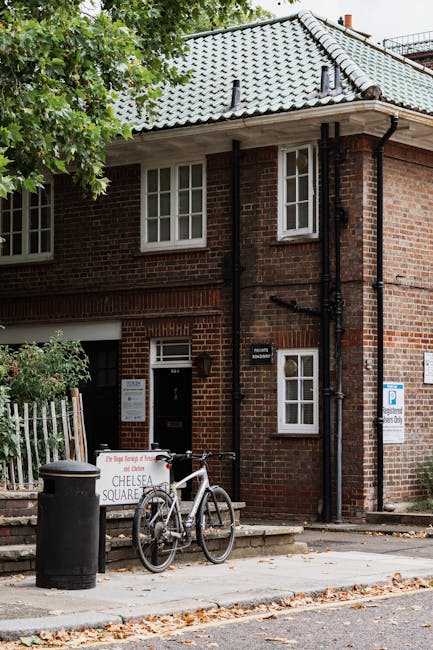 Exterior view of a brick residential building with a green-tiled pitched roof, featuring white-framed windows and a main entrance door. Outside, a white bicycle with a black saddle is parked against a small concrete step near a black trash bin. To the left of the bicycle, a white sign with red and black lettering indicates parking restrictions related to Chelsea Square, positioned on the sidewalk next to a metal fence with leafy green trees overhead providing partial shade. The scene captures a typical townhouse frontage during daytime, with natural light illuminating the brickwork and surroundings, and a visible parking sign on the building indicating parking permits and access for removals or home relocation logistics, consistent with Chelsea Harbour moving services provided by Movers Chelsea.