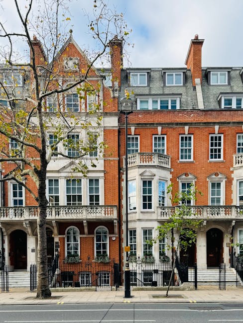 A row of historic red-brick and white-painted residential buildings with ornate architectural details line Sloane Square in Chelsea, London, during daylight. In the foreground, a paved sidewalk runs parallel to the street, with several leafless trees and a yellow and black roadside bollard. A black metal fence separates the pavement from the buildings' entrance steps, which lead up to front doors. The buildings feature large sash windows, some with decorative mouldings, and small balconies with stone balustrades. The scene suggests a residential area suitable for a home relocation or furniture transport, with no visible moving equipment or boxes present. The overall environment indicates an urban setting prepared for a professional house removal service like Movers Chelsea, with natural light illuminating the facades against a partly cloudy sky.