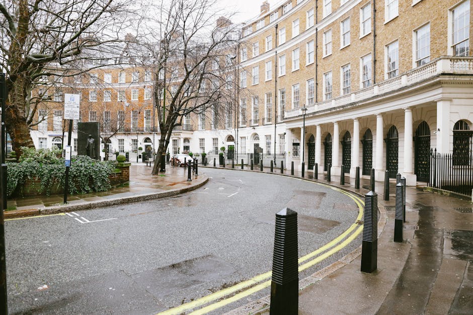 A row of historic red-brick and white-painted residential buildings with ornate architectural details line Sloane Square in Chelsea, London, during daylight. In the foreground, a paved sidewalk runs parallel to the street, with several leafless trees and a yellow and black roadside bollard. A black metal fence separates the pavement from the buildings' entrance steps, which lead up to front doors. The buildings feature large sash windows, some with decorative mouldings, and small balconies with stone balustrades. The scene suggests a residential area suitable for a home relocation or furniture transport, with no visible moving equipment or boxes present. The overall environment indicates an urban setting prepared for a professional house removal service like Movers Chelsea, with natural light illuminating the facades against a partly cloudy sky.